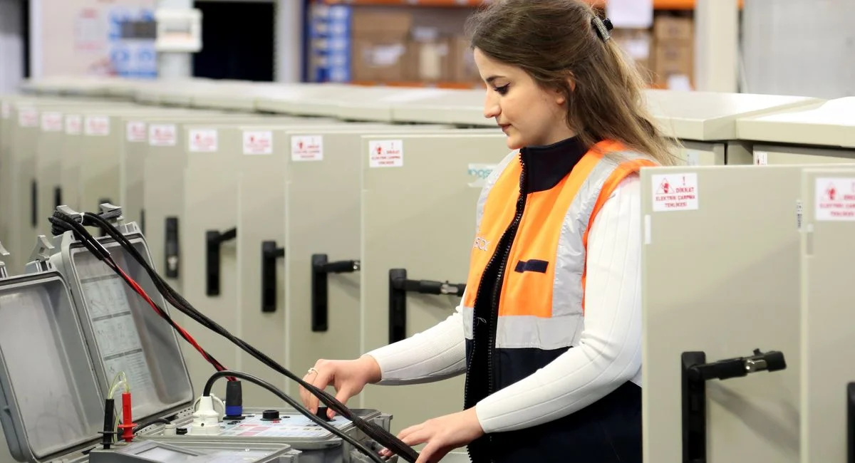 A female electrician in safety gear checks electrical panels indoors.
