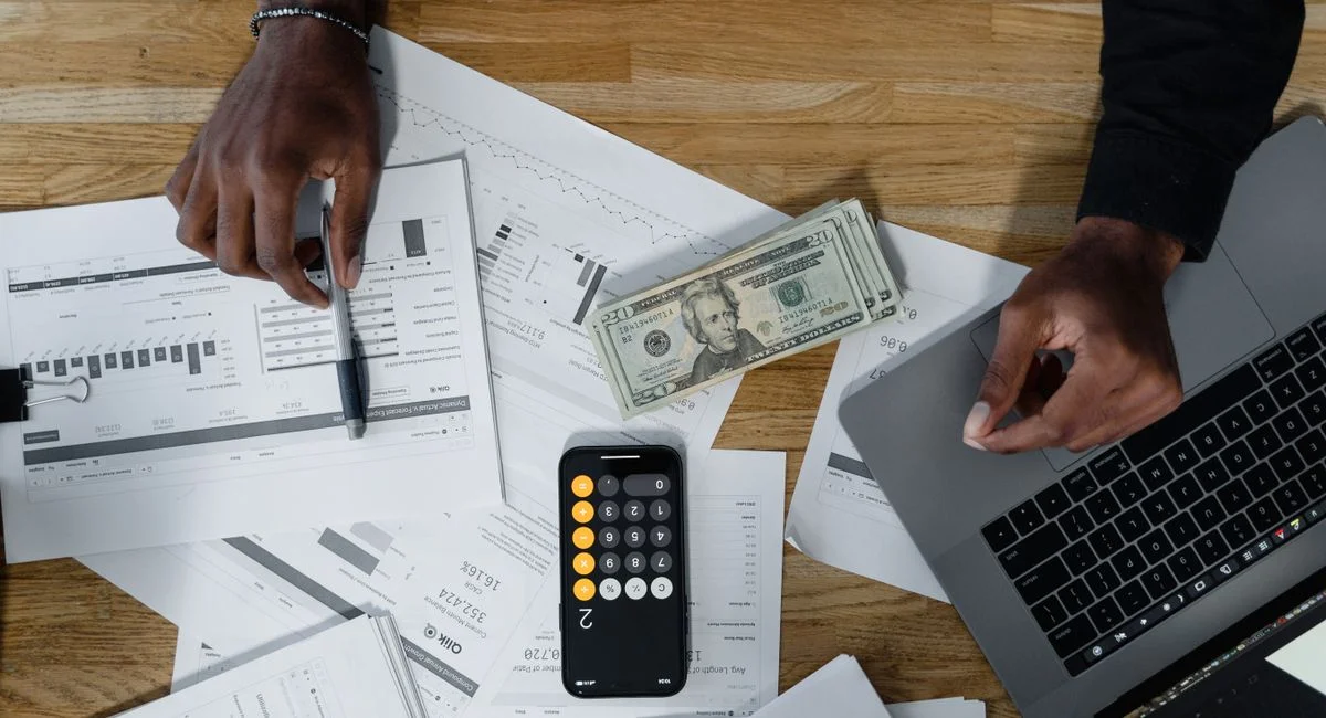 Hands calculate finances with papers, cash, and a laptop on a wooden desk.