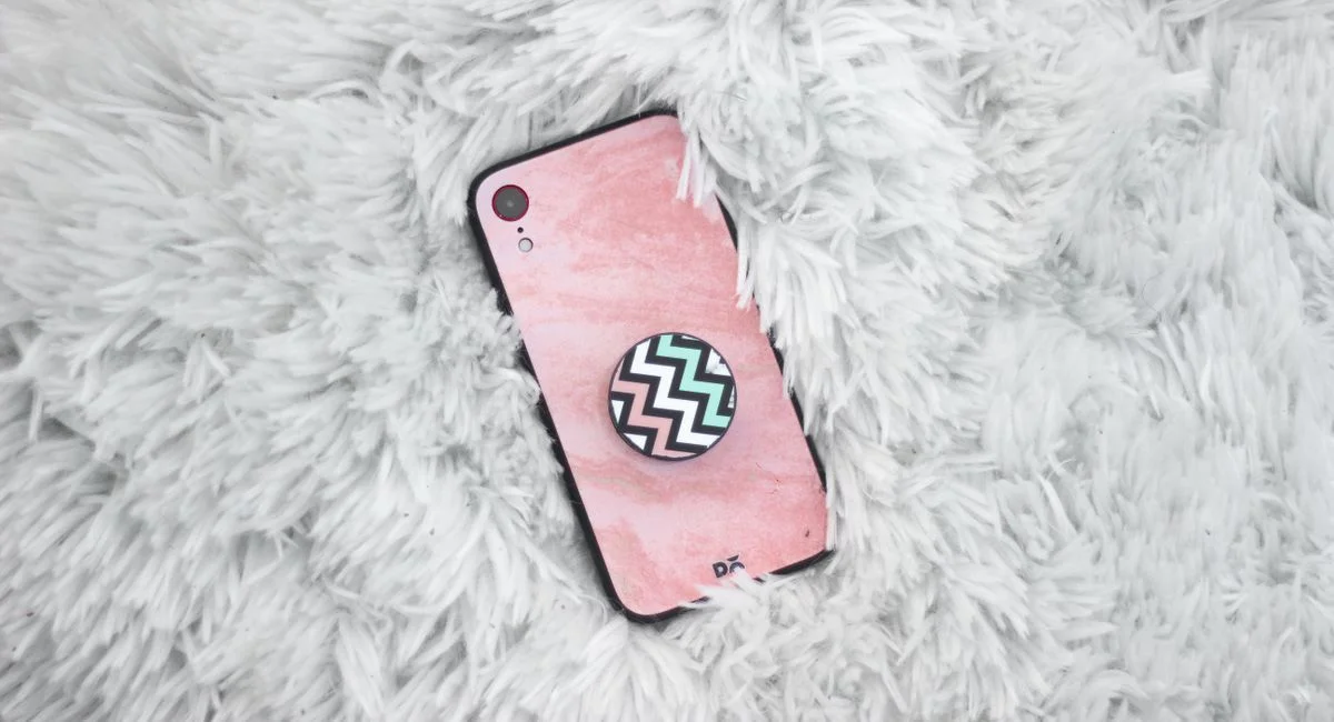 Top view of a mobile phone with a pink case and zigzag pop socket on a fluffy white rug.